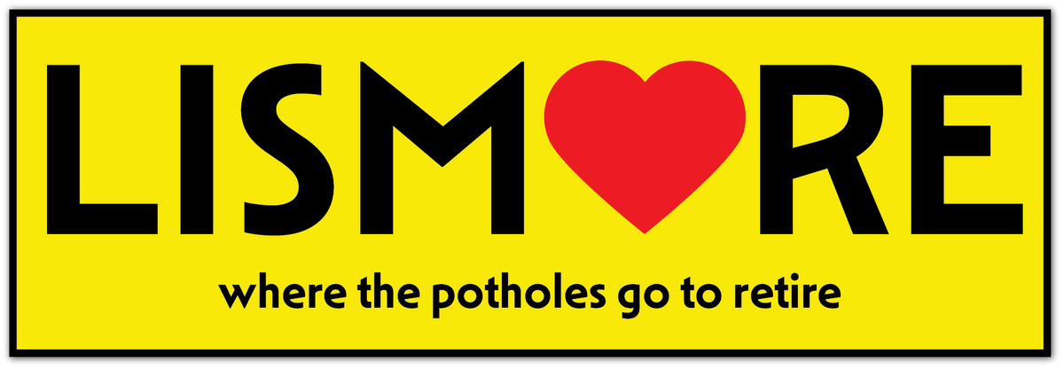 A bright yellow rectangular sign or sticker with a black border. The word 'LISMORE' is printed in large black letters, with a red heart replacing the 'O'.
